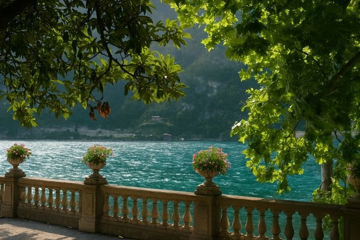 A stone terrace with flower urns under green branches, looking out over a bright turquoise lake.